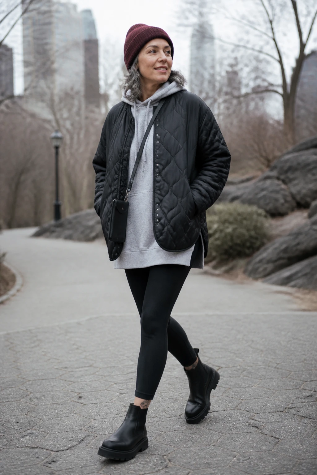 A stylish mid-50s woman wears a black quilted liner jacket with a long hoodie, opaque leggings, and sleek Chelsea boots with a burgundy beanie.