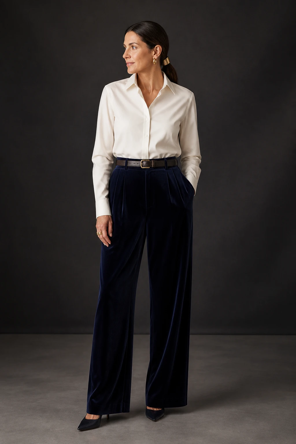 An elegant woman in her late 40s wears velvet wide-leg trousers with a tucked ivory poplin shirt, pointed pumps, and matte gold jewelry.