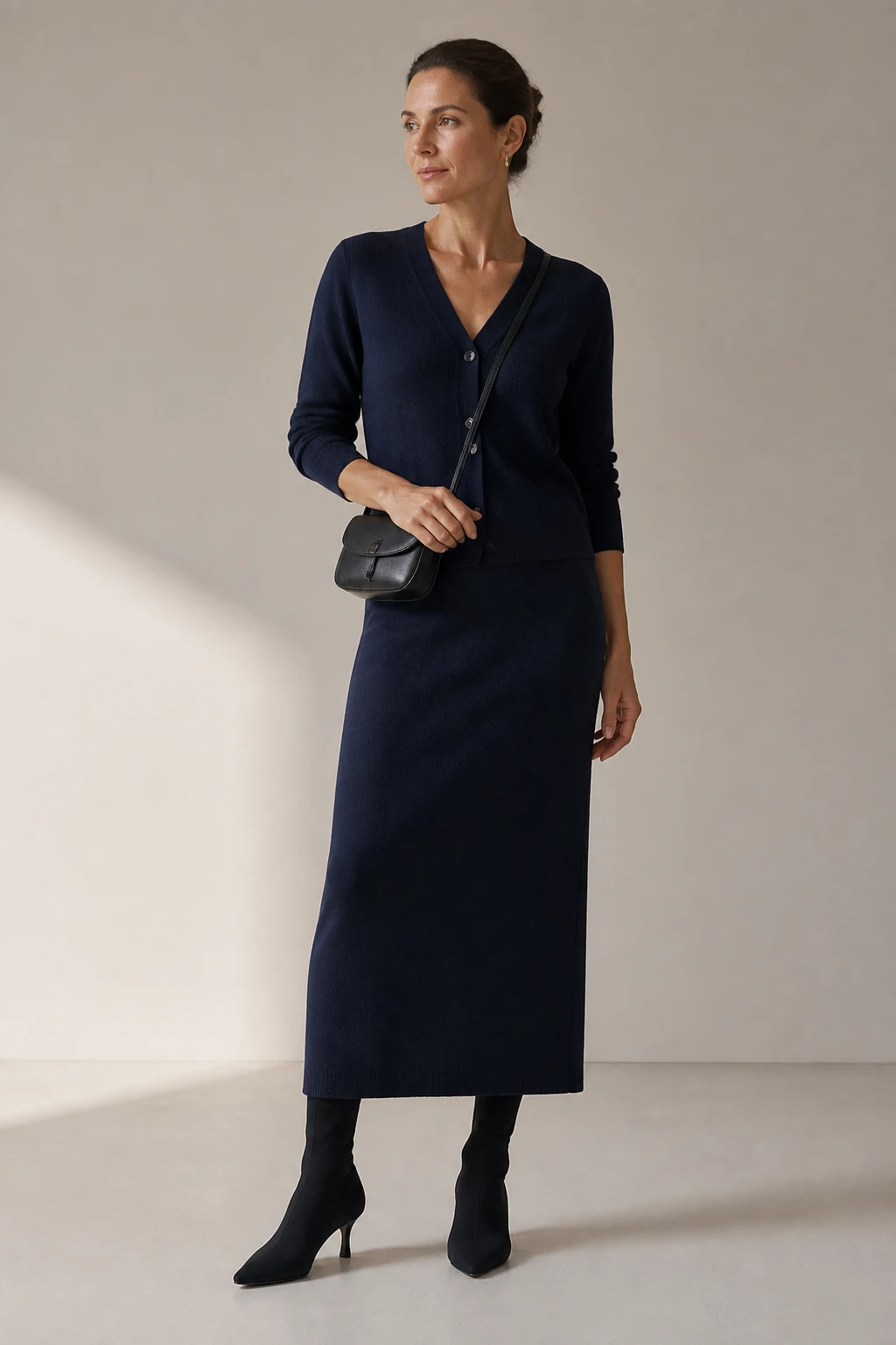 A sophisticated mid-40s woman wears a deep navy fine-gauge cardigan and matching column skirt with slim sock boots and a compact crossbody for a sleek monochrome look.