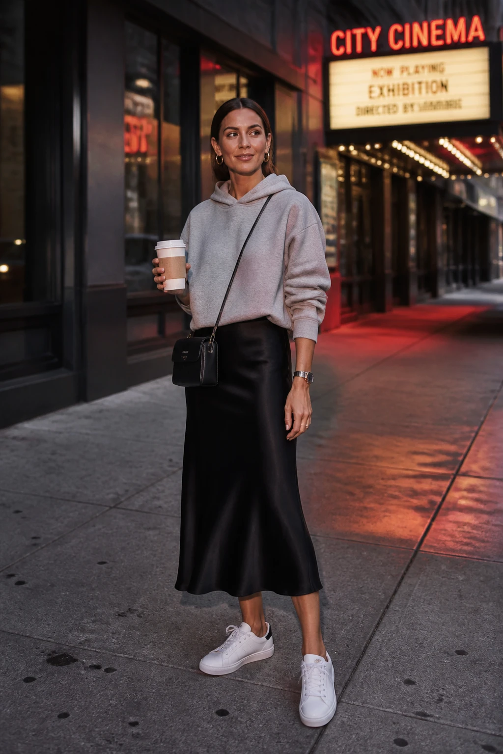 A sophisticated mid-30s woman styles a black satin midi skirt with a heather-gray cropped hoodie and white court sneakers.
