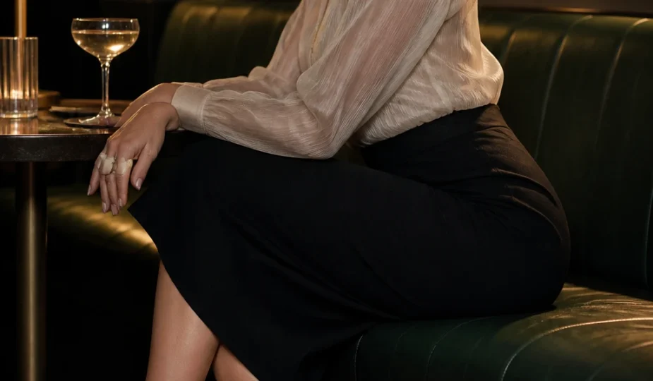 A sophisticated late-30s woman wears a lined sheer organza blouse with a high-waist pencil skirt and slingbacks, finished with pearl studs.
