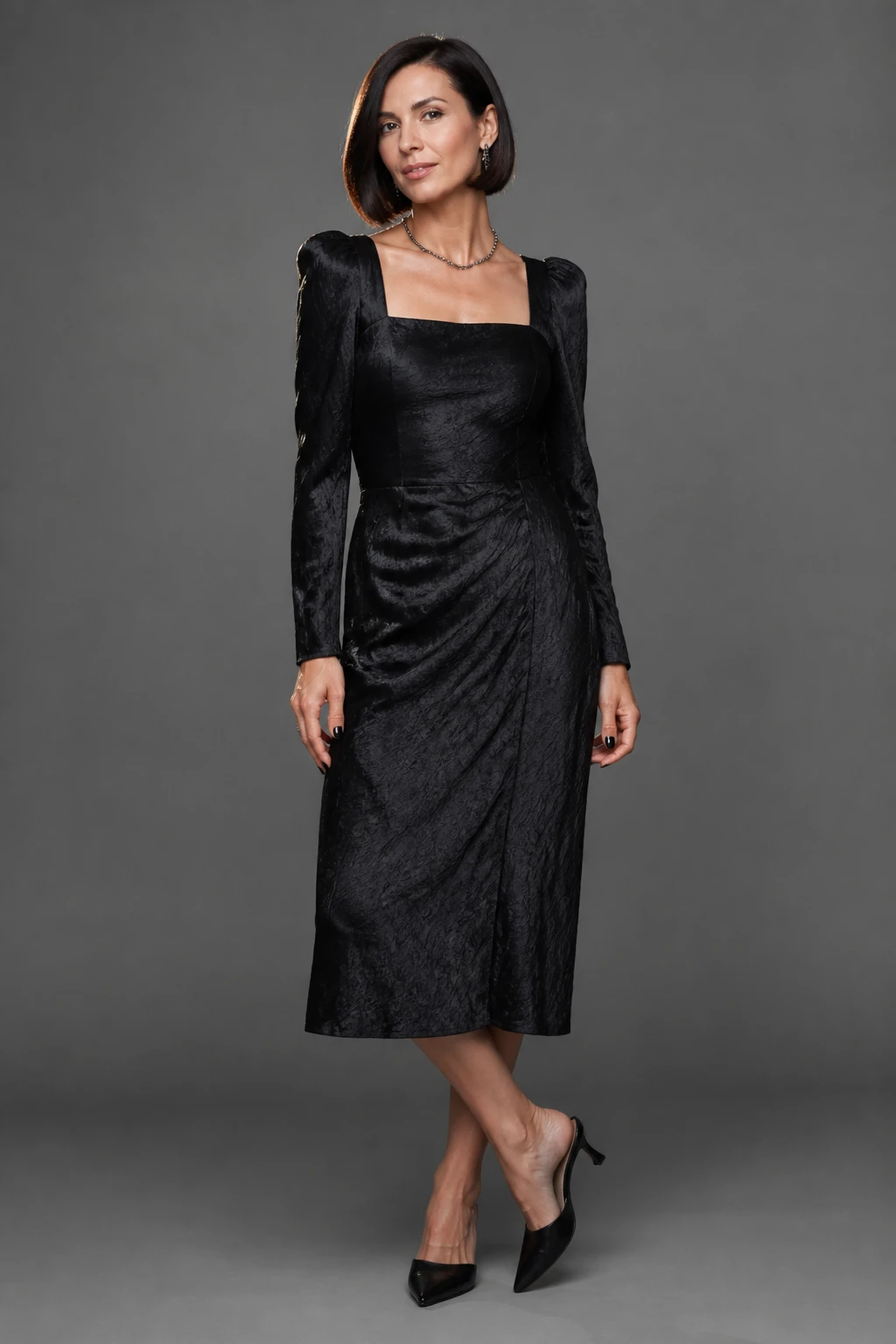 A poised woman in her mid-30s wears a textured black midi dress with square neckline, black kitten mules, and hematite-toned jewelry in studio light.