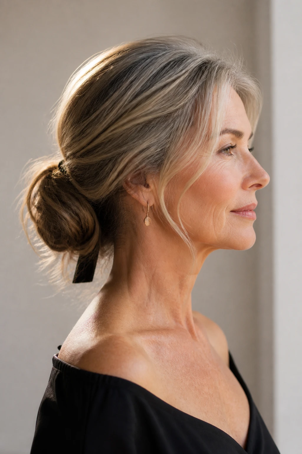 An elegant mid-50s woman wears a low soft bun with two wispy face pieces that trace along her glasses.