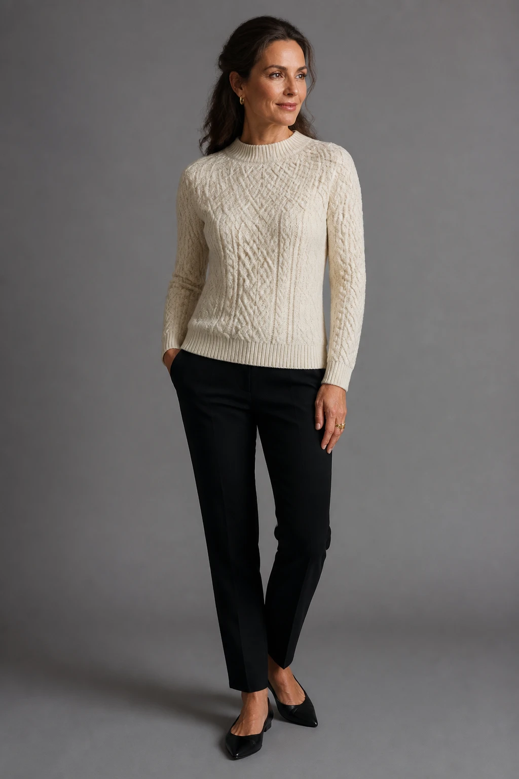An elegant mid-40s woman balances an ecru fisherman sweater with lean black trousers and pointed flats, minimal jewelry, and a half-up hairstyle.